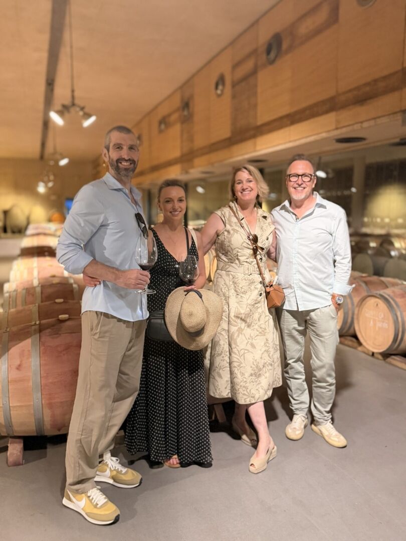 IMG_3463 - Petsolt Family Four friends smiling and posing with wine glasses in a winery, barrels in the background.