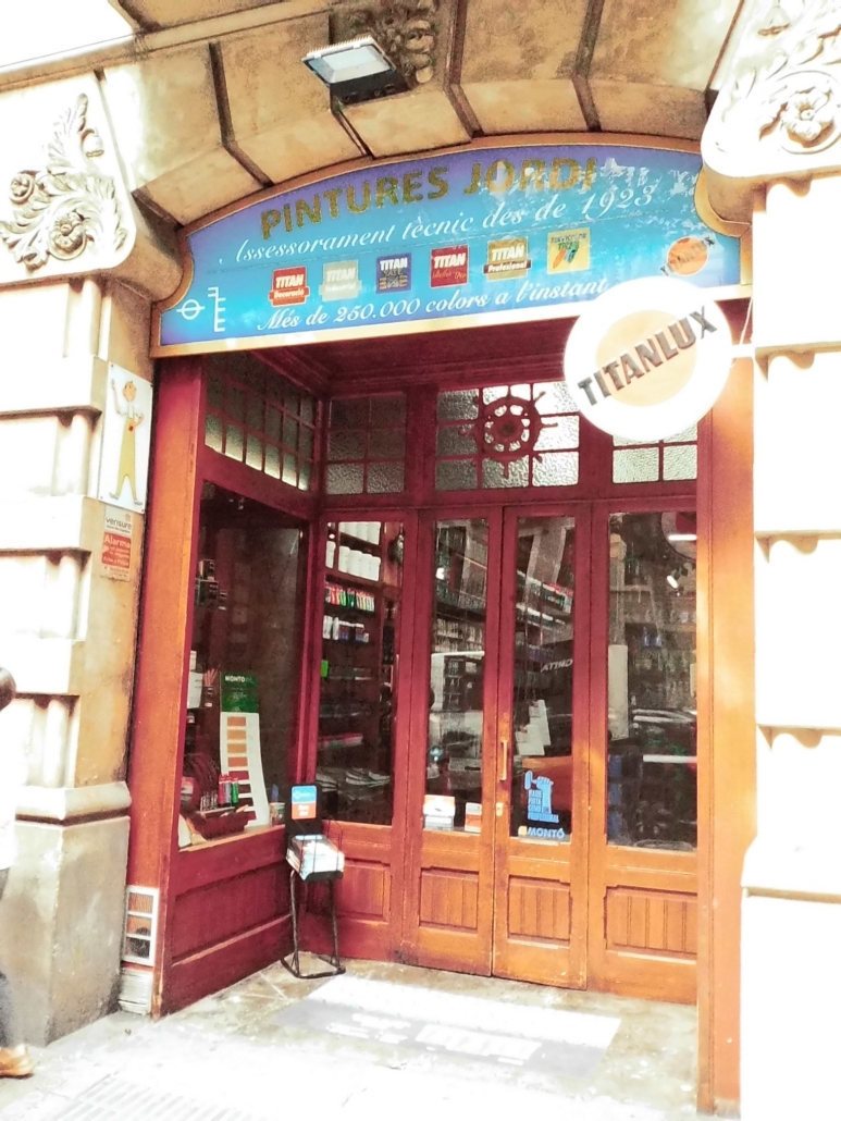 Block - Left - 15 of 16 - Petsolt Family Front of a paint shop with a blue sign reading 'PINTURES JORDI' and brand logos, glass double doors, and shelves of products visible inside.