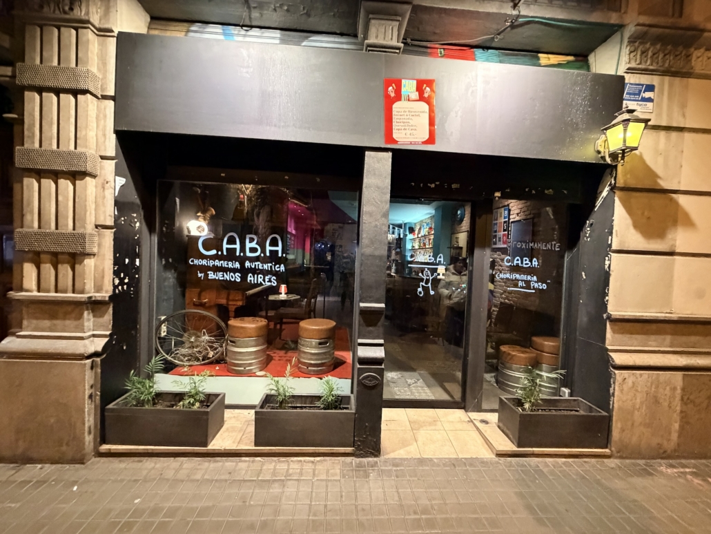 Block - Left - 8 of 16 - Petsolt Family Night view of a black storefront for C.A.B.A. Choripería, with glass doors, street barrels and rectangular planters outside, and stone building façades on both sides.