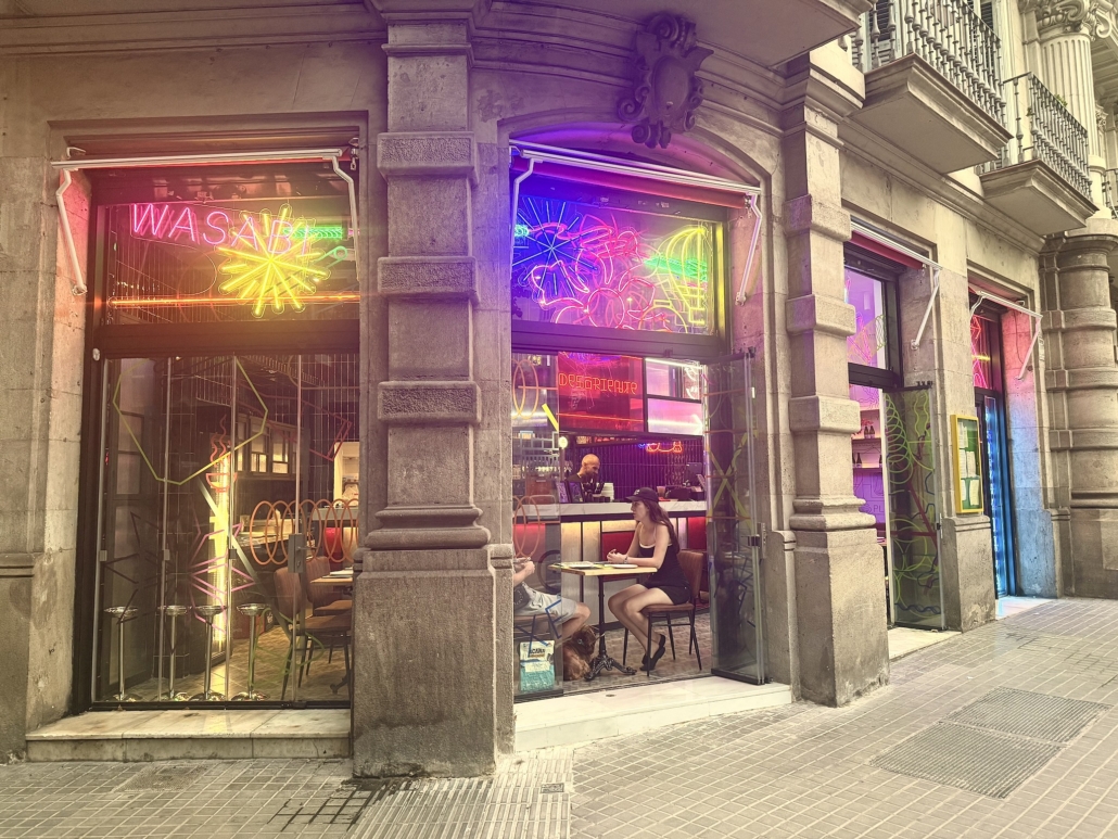 Neon-lit cafe storefront on a stone building, two people seated at a small table outside, colorful neon signs in the windows.