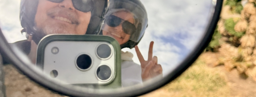 Person wearing helmet and sunglasses reflected in a round mirror, smiling at the camera outdoors, making a peace sign with fingers