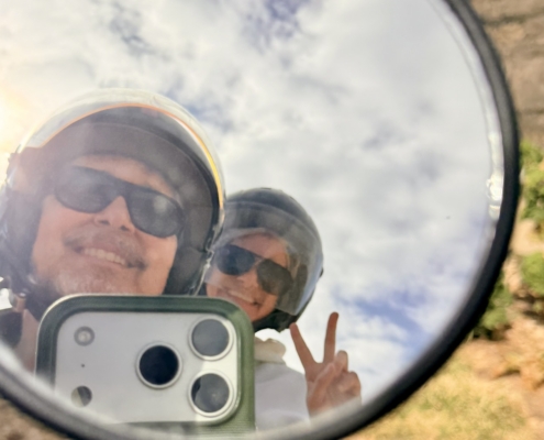 Person wearing helmet and sunglasses reflected in a round mirror, smiling at the camera outdoors, making a peace sign with fingers