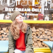 Woman in a beanie and green jacket sits by a yellow bench outside a deli, looking upward; window reads 'Day Dream Deli' with white graphics and stacked wood behind.