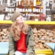 Woman in a beanie and green jacket sits by a yellow bench outside a deli, looking upward; window reads 'Day Dream Deli' with white graphics and stacked wood behind.