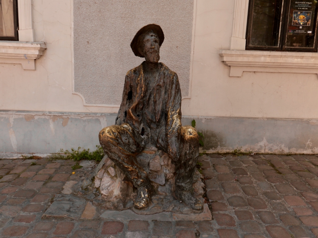 Weathered bronze statue of a seated figure wearing a hat, set against a pale building with windows and a cobblestone ground.