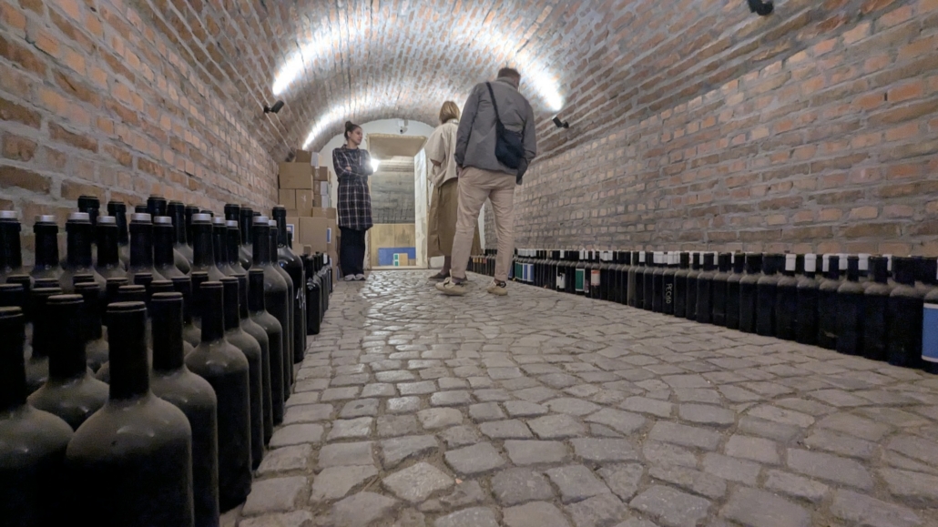 Brick-walled wine cellar with arched ceiling and long rows of bottles along both walls; three people inspect boxes in the back.