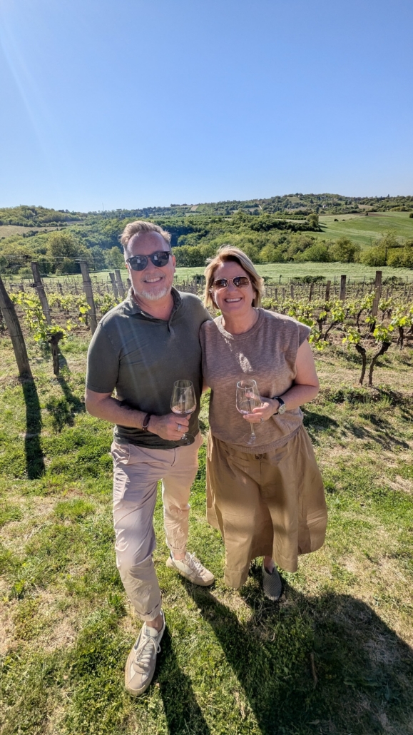 Smiling couple wearing sunglasses in a vineyard, each holding a wine glass on a sunny day with rows of grapevines behind them.