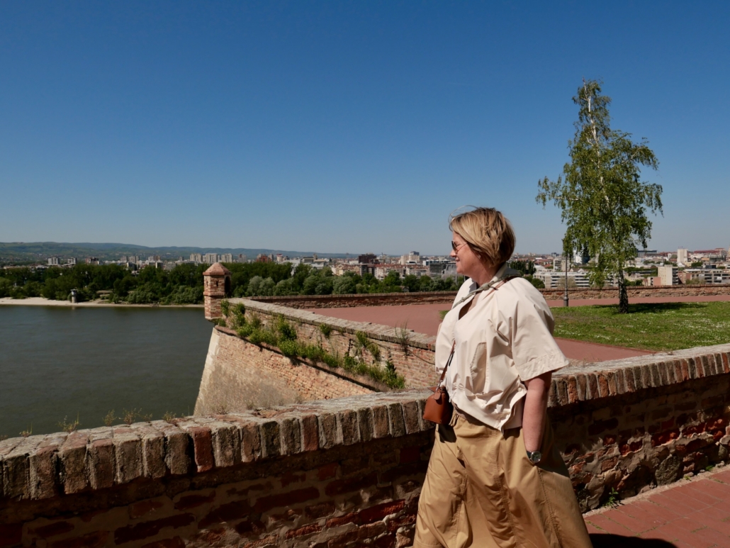 Woman in a light beige trench coat walking along a brick riverfront wall with a city skyline and a tall tree in the background.