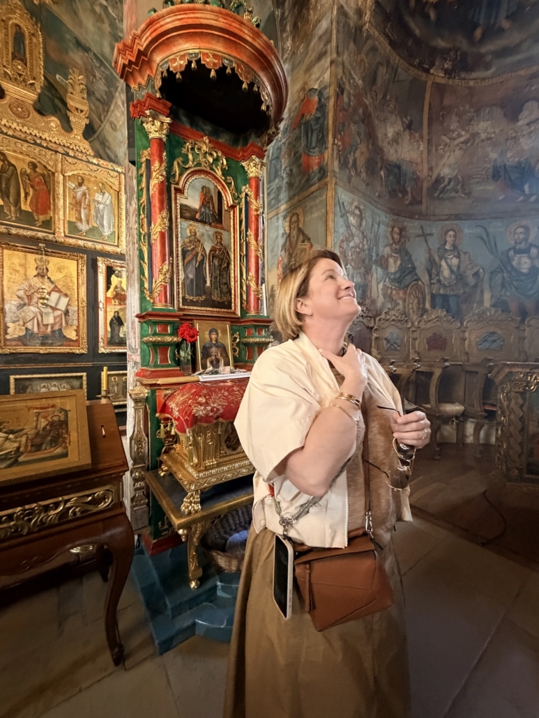Woman in a beige coat stands beside an ornate altar in a richly painted church interior, looking up with a smile.