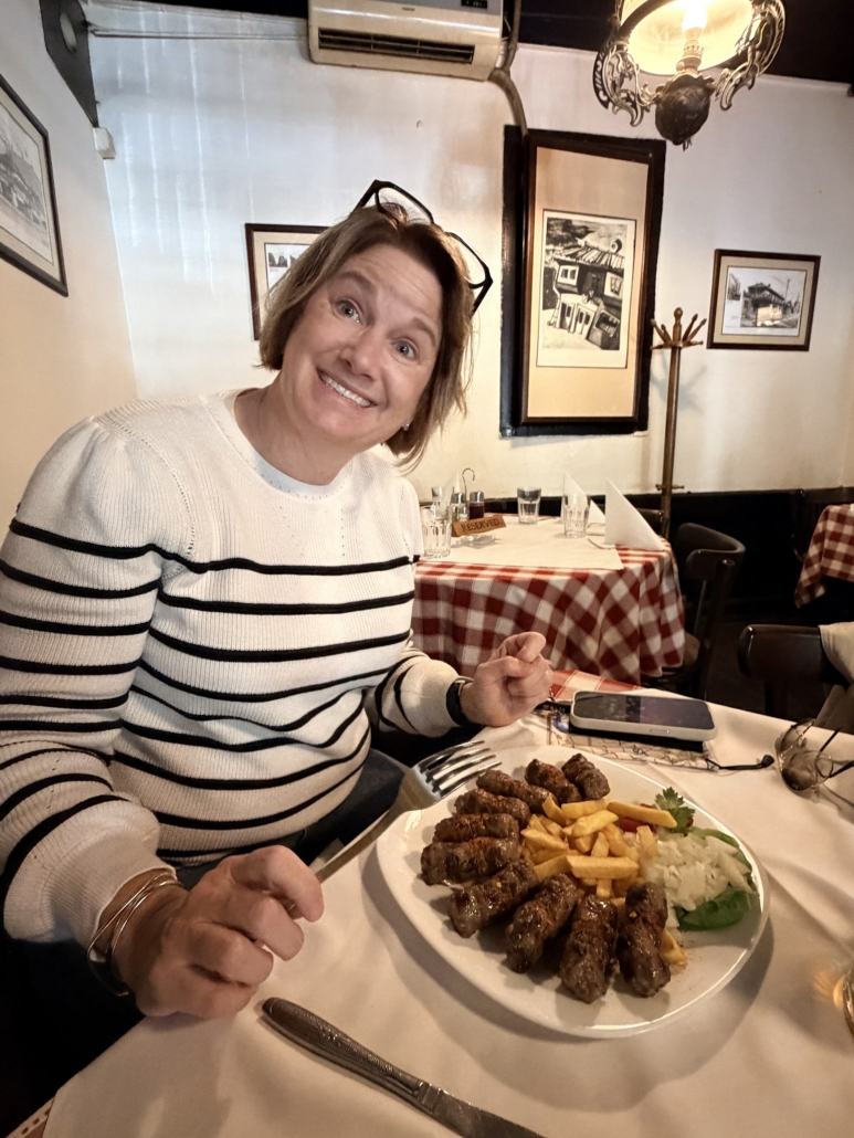 Smiling woman in a striped sweater sits at a table with a plate of grilled meat and fries, in a cozy restaurant setting.