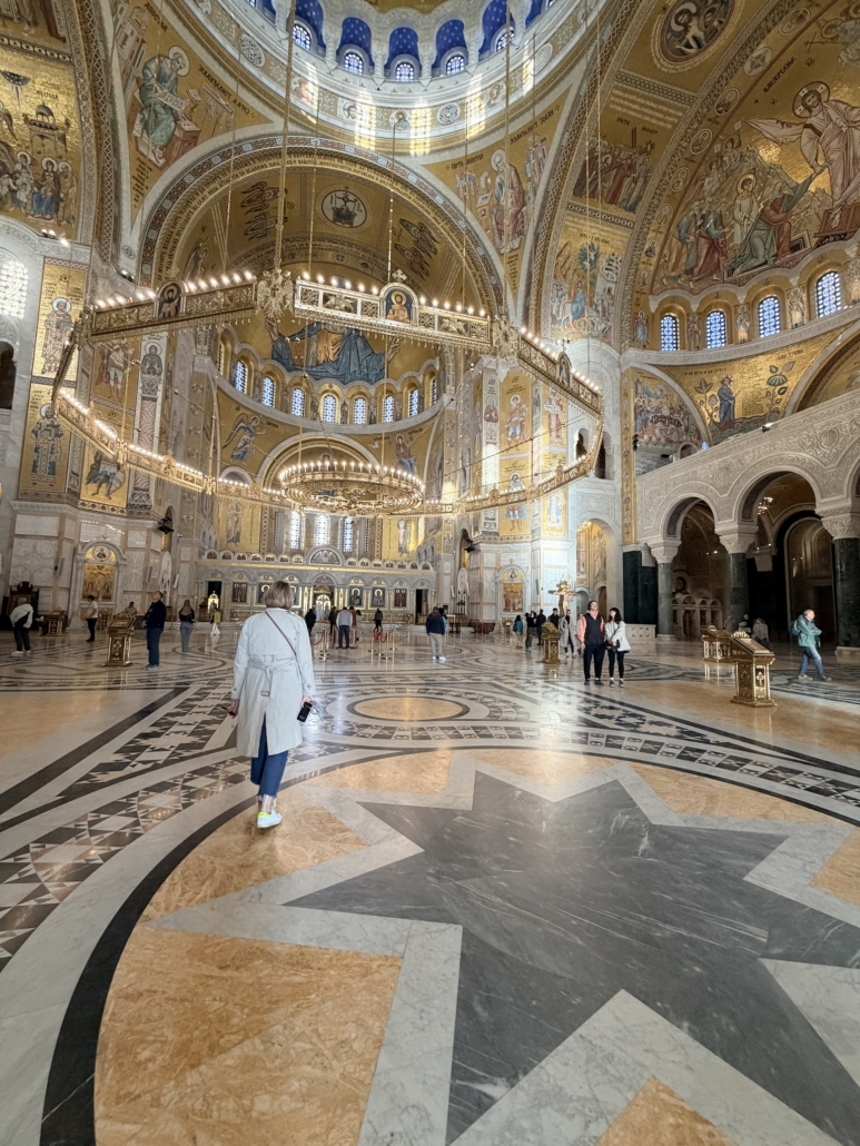 Interior of a grand Orthodox church with gold mosaics, chandeliers, and arched ceilings; visitors walk on a marble floor with geometric patterns.