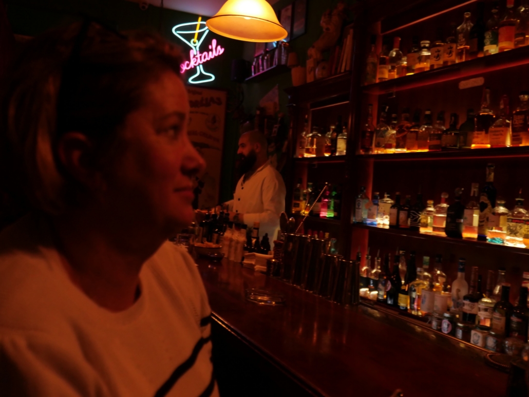 Dimly lit bar scene with a neon cocktail sign, shelves of liquor, and a bartender behind the counter; foreground person in profile.