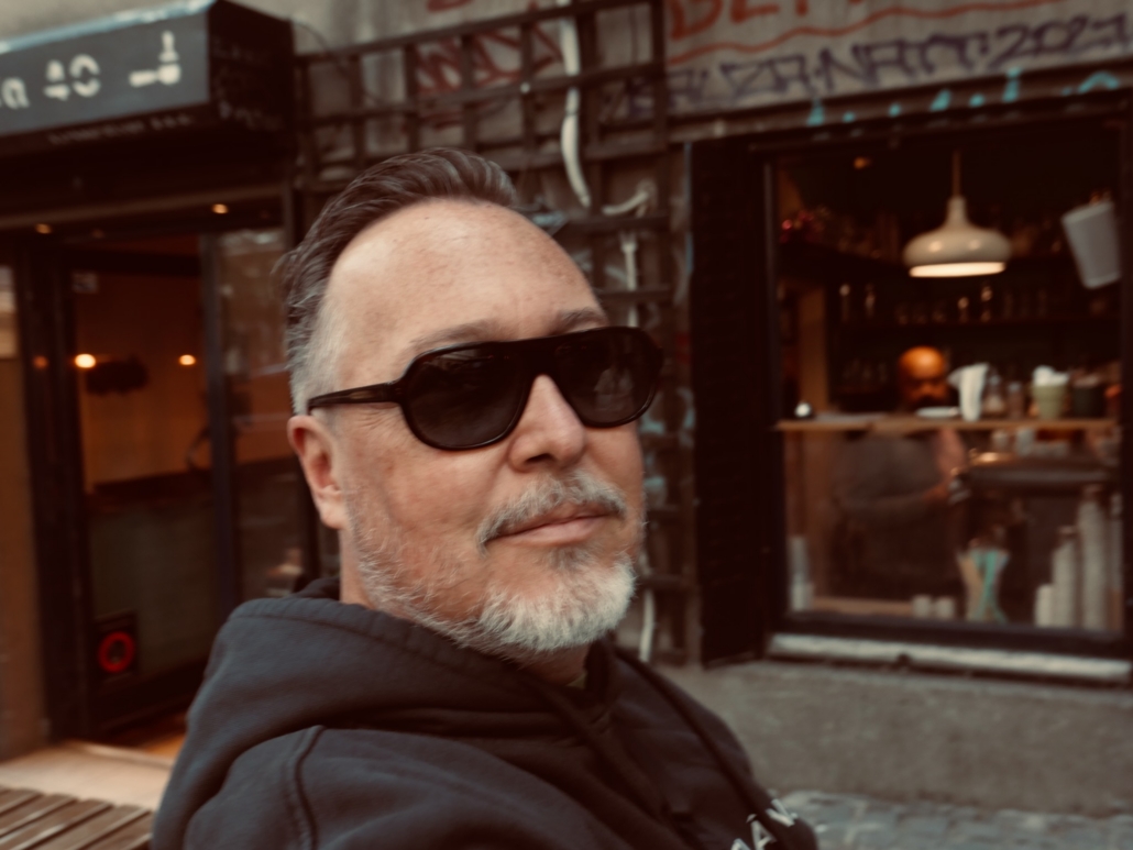 Close-up of a man with a gray beard wearing dark sunglasses, seated outdoors in front of a cafe storefront.