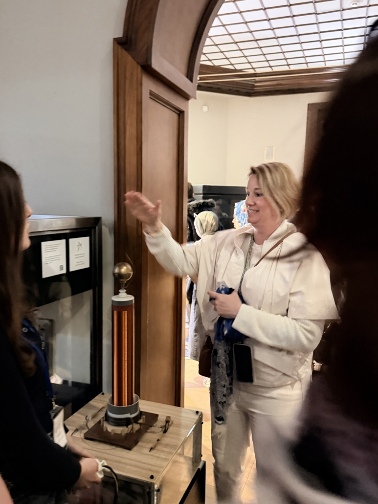 Smiling woman in a white jacket points at a tall copper instrument in a glass display case inside a museum setting.