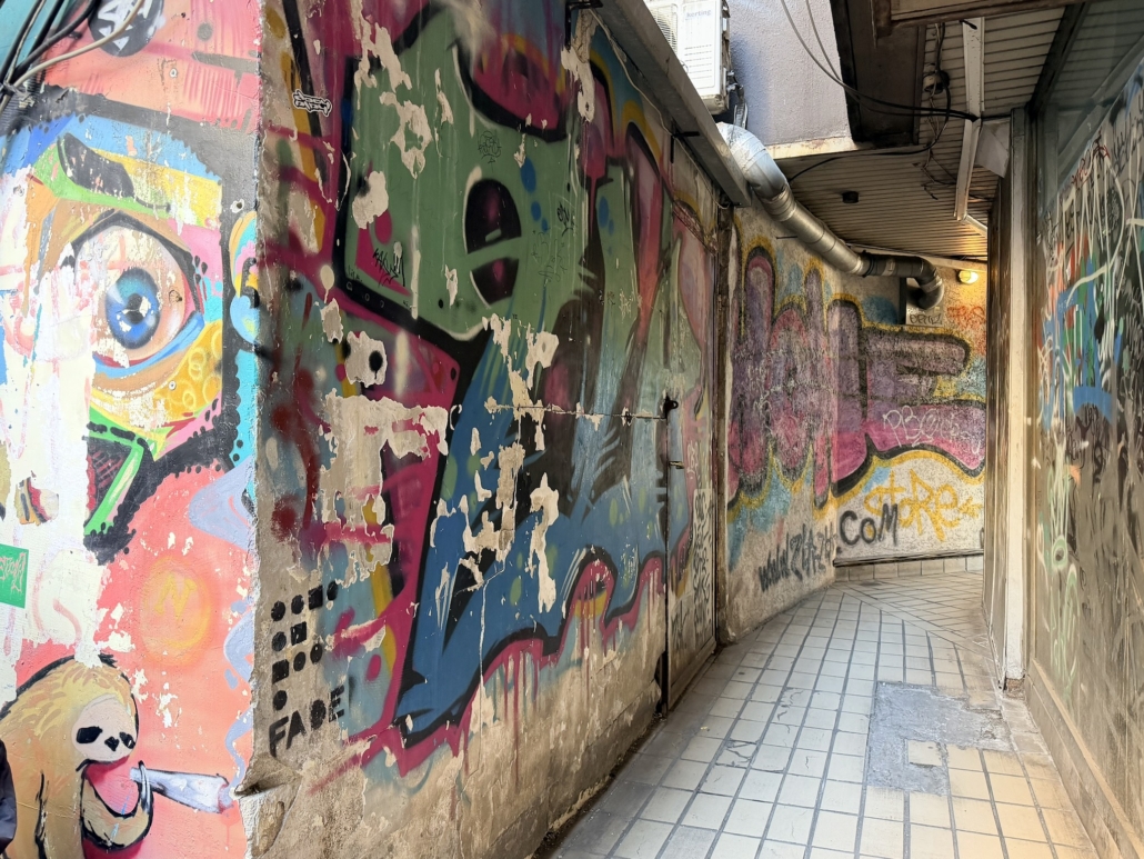 Narrow alleyway lined with colorful graffiti murals, peeling paint, and graffiti-covered walls, with tiled ground and overhead pipes leading to a lighted end.