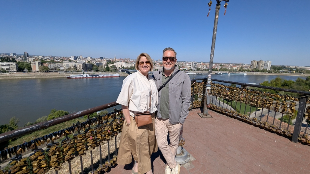 Smiling couple standing on a railing covered with love locks, overlooking a river and city skyline on a sunny day.