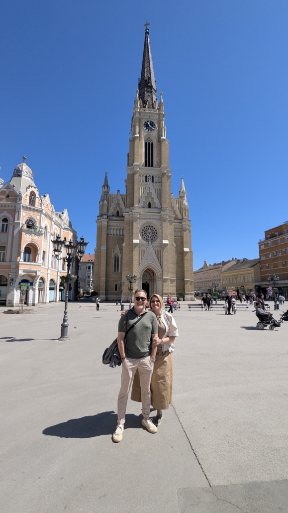Couple posing in a plaza in front of a tall Gothic church with a clock tower under a clear blue sky there are pedestrians around.