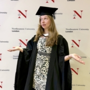 Graduate in cap and gown gesturing with hands in front of a Northeastern University London backdrop.