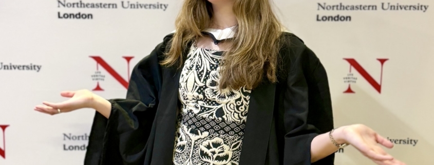 Graduate in cap and gown gesturing with hands in front of a Northeastern University London backdrop.