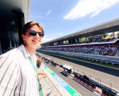 2026-08- Budapest Cams 18th - 11 of 11 - Petsolt Family - Petsolt Family Selfie of a smiling person wearing sunglasses at a large outdoor race track with a crowded stadium and blue sky.