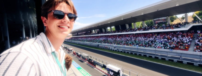Selfie of a smiling person wearing sunglasses at a large outdoor race track with a crowded stadium and blue sky.
