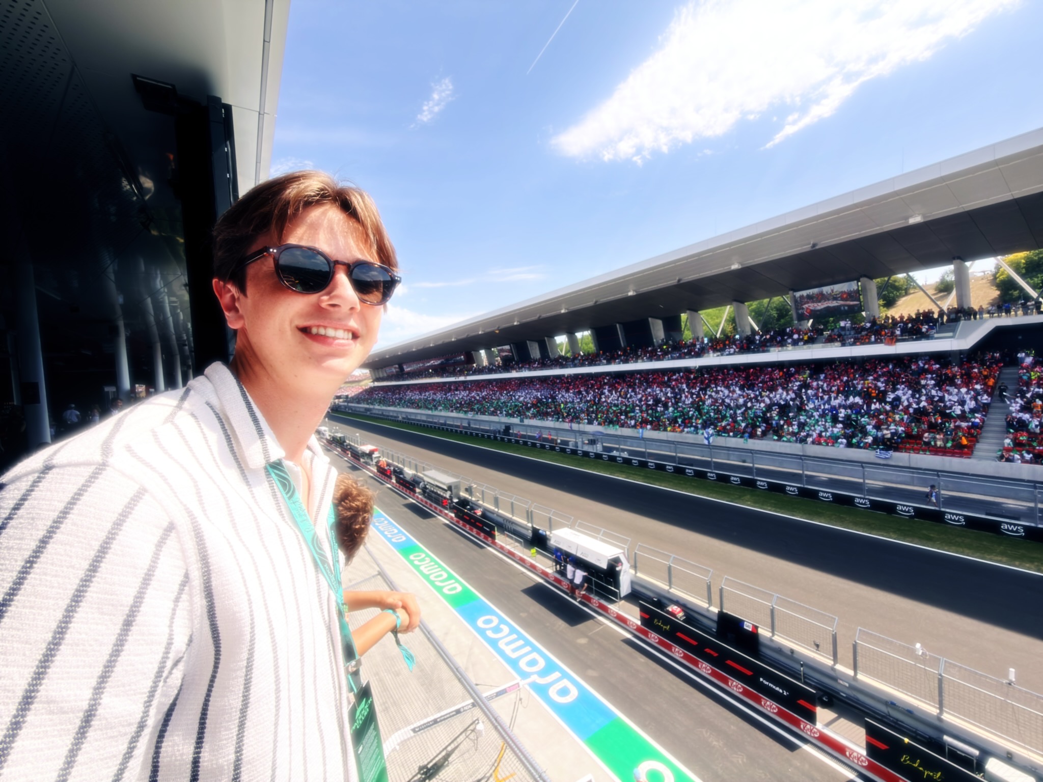 Selfie of a smiling person wearing sunglasses at a large outdoor race track with a crowded stadium and blue sky.