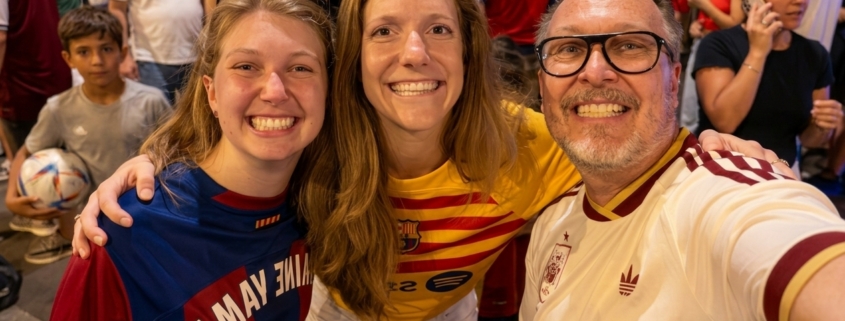 Three friends take a selfie in a crowded street crowd, all smiling and wearing colorful sports jerseys.