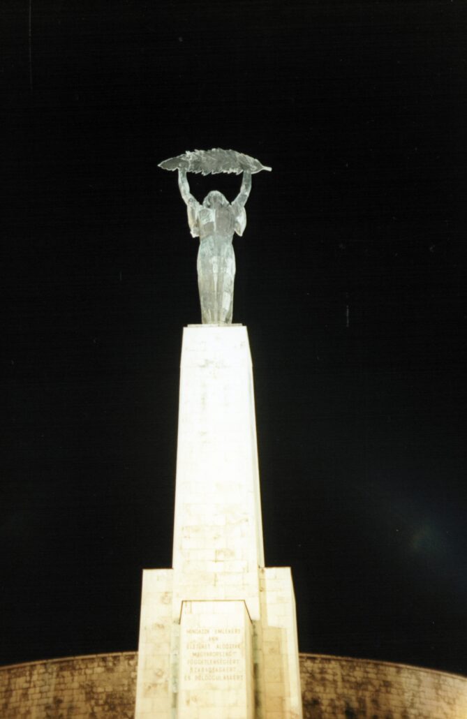 00394_p_w16aneafypk0394 - Petsolt Family Night view of a tall monument: a statue atop an obelisk lifting a leafy branch above its head, illuminated against a dark sky.