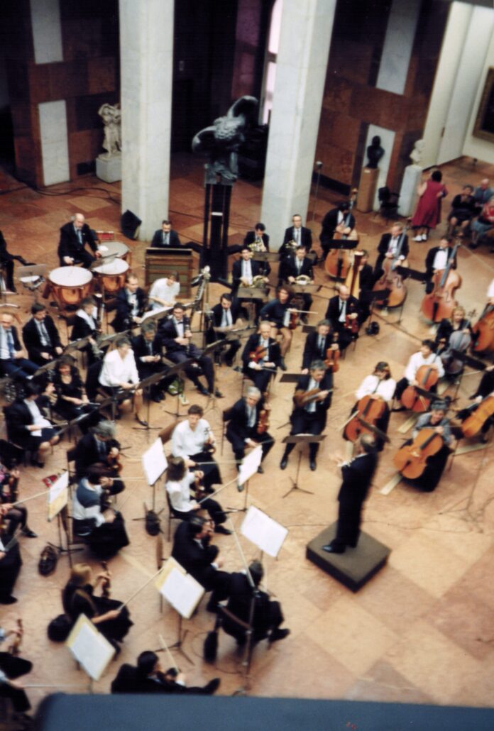 00401_p_w16aneafypk0401 - Petsolt Family Conductor leads a full orchestra on a wooden stage in a large hall, with musicians and instruments arranged around him.