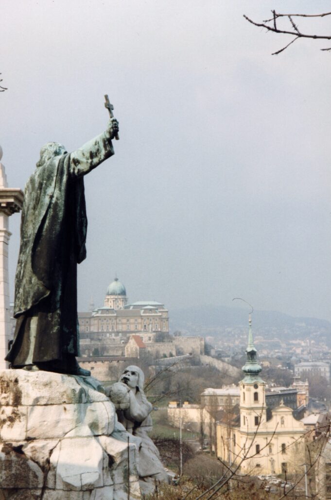 00402_p_w16aneafypk0402 - Petsolt Family Bronze statue in a robe raises a cross above a stone pedestal, overlooking a historic city with domed buildings in the distance