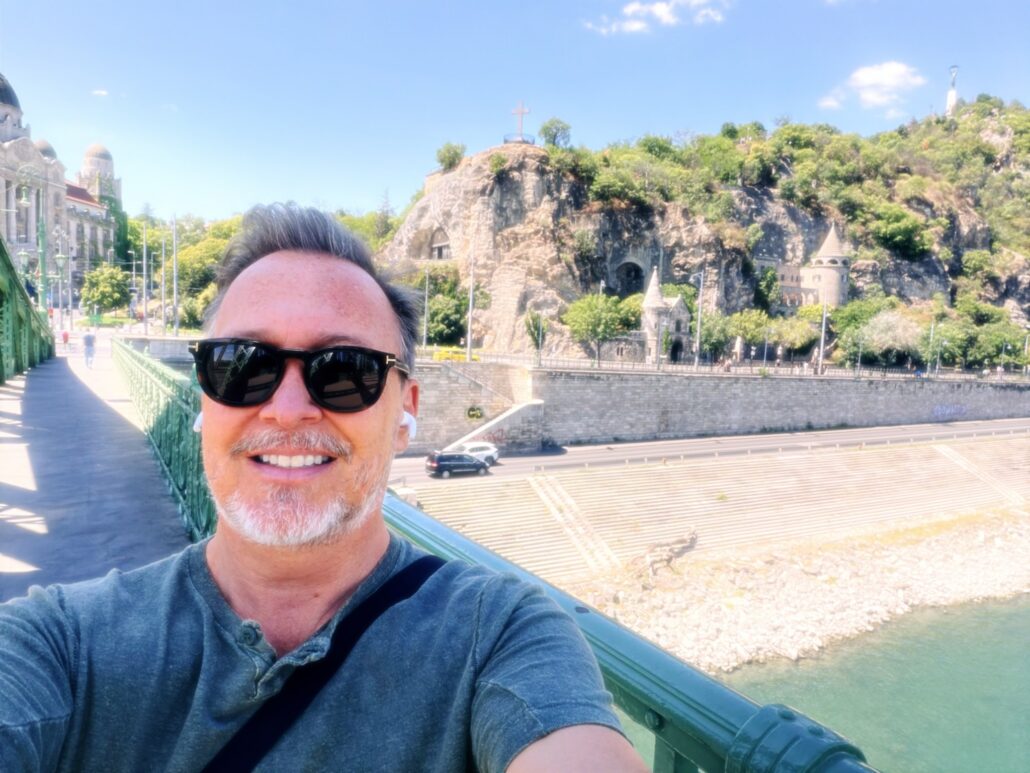 2026-08- Budapest Cams 18th - 2 of 8 - Petsolt Family Selfie of a man wearing sunglasses on a green railing bridge, with a river and hillside featuring a cross in the background.