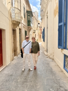Two adults stand in a sunlit narrow Mediterranean alley with stone buildings and blue window shutters. (informative)
