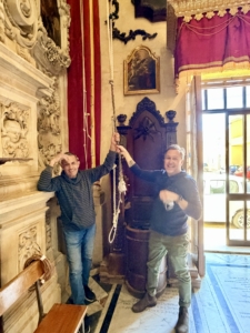 Two smiling men hold a long rope in a richly decorated room with ornate stone carvings and red velvet drapes nearby.
