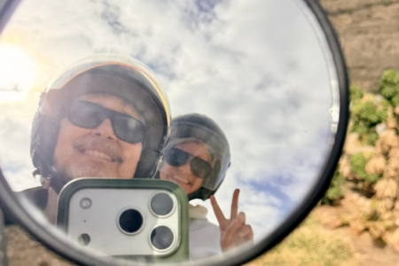 Person wearing helmet and sunglasses reflected in a round mirror, smiling at the camera outdoors, making a peace sign with fingers