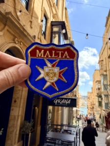 Patch reading 'MALTA' with a Maltese cross emblem held up against a sunny Maltese street scene.