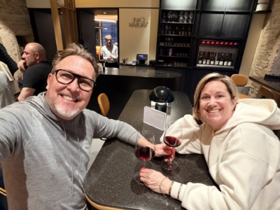 Two friends smiling for a selfie at a bar, holding glasses of red wine with a bartender and wine shelves in the background]