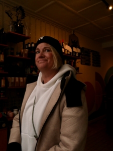 Woman in a beige coat and white scarf stands in a dimly lit store, shelves behind her filled with bottles and jars.