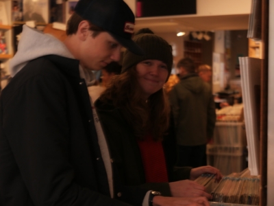 Two people leaning over a keyboard, one wearing a black cap and hoodie and the other a knit hat, playing together in a cozy shop.