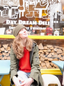 Woman in a beanie and green jacket sits by a yellow bench outside a deli, looking upward; window reads 'Day Dream Deli' with white graphics and stacked wood behind.