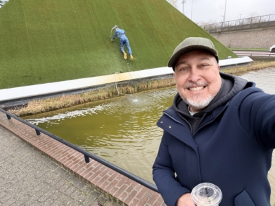 Stopping the leak & saving the day Smiling man in a navy jacket and flat cap taking a selfie beside a large green grassy hill sculpture by a canal edge.