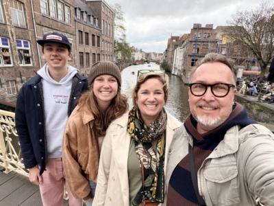 Four friends posing for a selfie on a canal bridge with brick buildings and boats in the background.