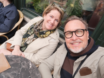 Couple taking a cheerful selfie at an outdoor cafe, with a marble table in the foreground and greenery in the background.