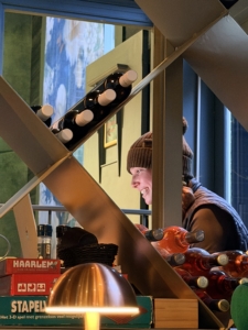 Working along the way Wine bottles lined along a slanted display rack in a shop, with a smiling person wearing a brown knit hat in the background.