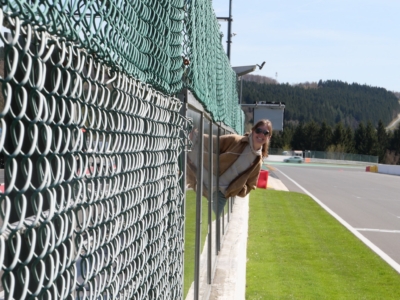 Person leaning on a glass barrier beside a tall green chain-link fence at a race track on a sunny day, with trees in the background.