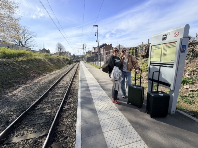 The big train station in Spa Two women standing on a train platform with rolling suitcases, smiling at the camera as tracks extend into the distance under a blue sky.