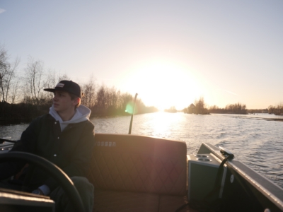 Our captain Young man wearing a cap and jacket sits in a boat on a calm river at sunset, with trees along the bank behind him.