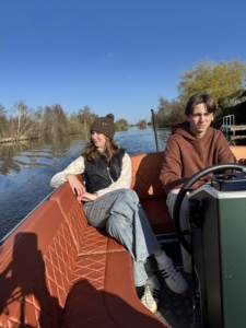 Two people sit on an orange cushioned boat along a calm canal with a clear blue sky above.