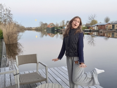 Girl standing on a wooden dock by a calm lake, mouth open in surprise, wearing a black quilted vest and gray sweatpants, with houses in the background.