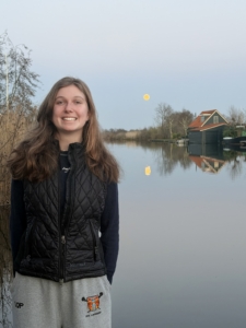Smiling young woman in a black quilted vest standing by a calm river with a house in the background under a pale sky.
