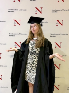 Graduate in cap and gown gesturing with hands in front of a Northeastern University London backdrop.